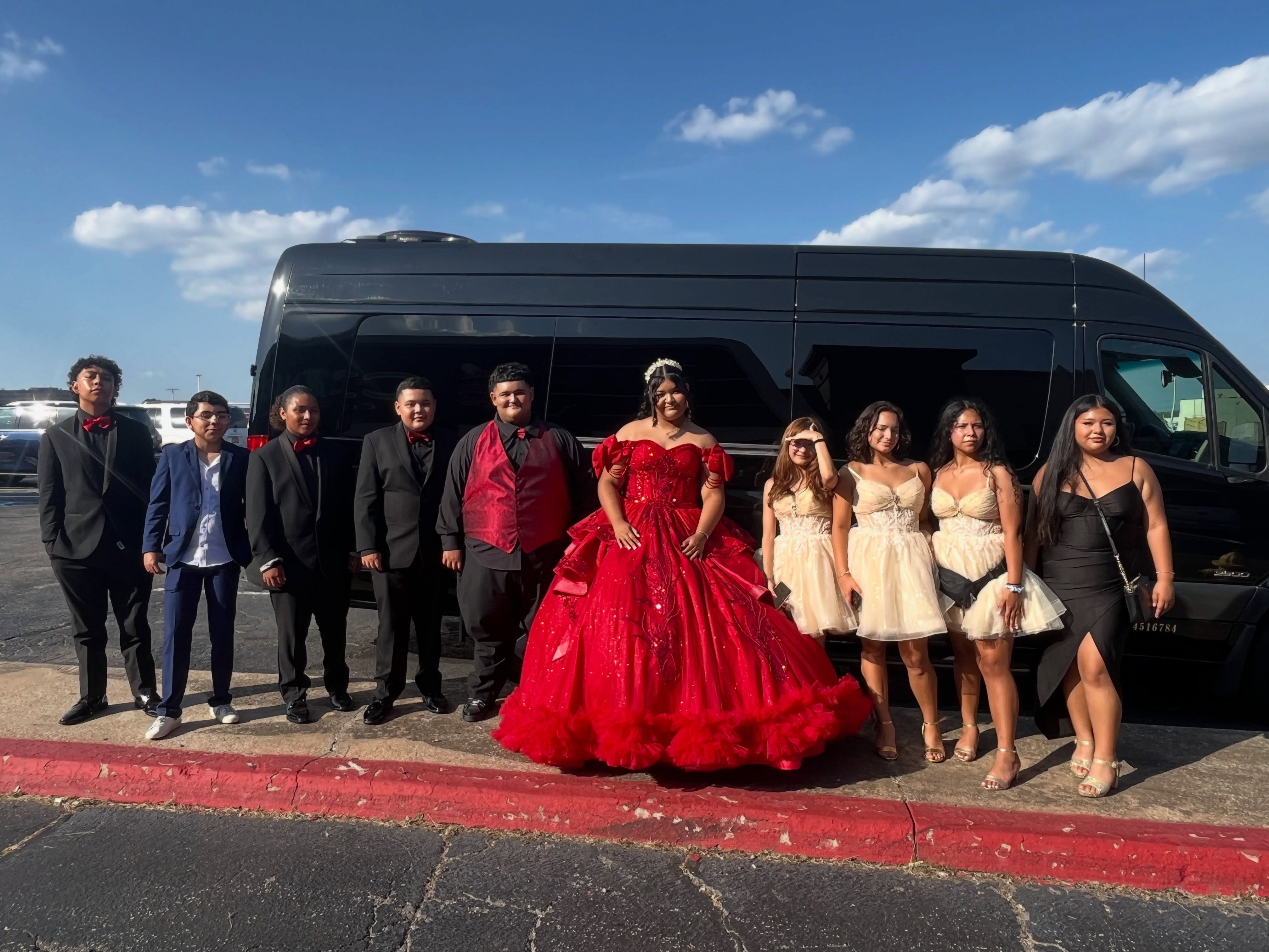A group of teens in formal attire pose in front of a black Mercedes Sprinter van in a parking lot under blue sky