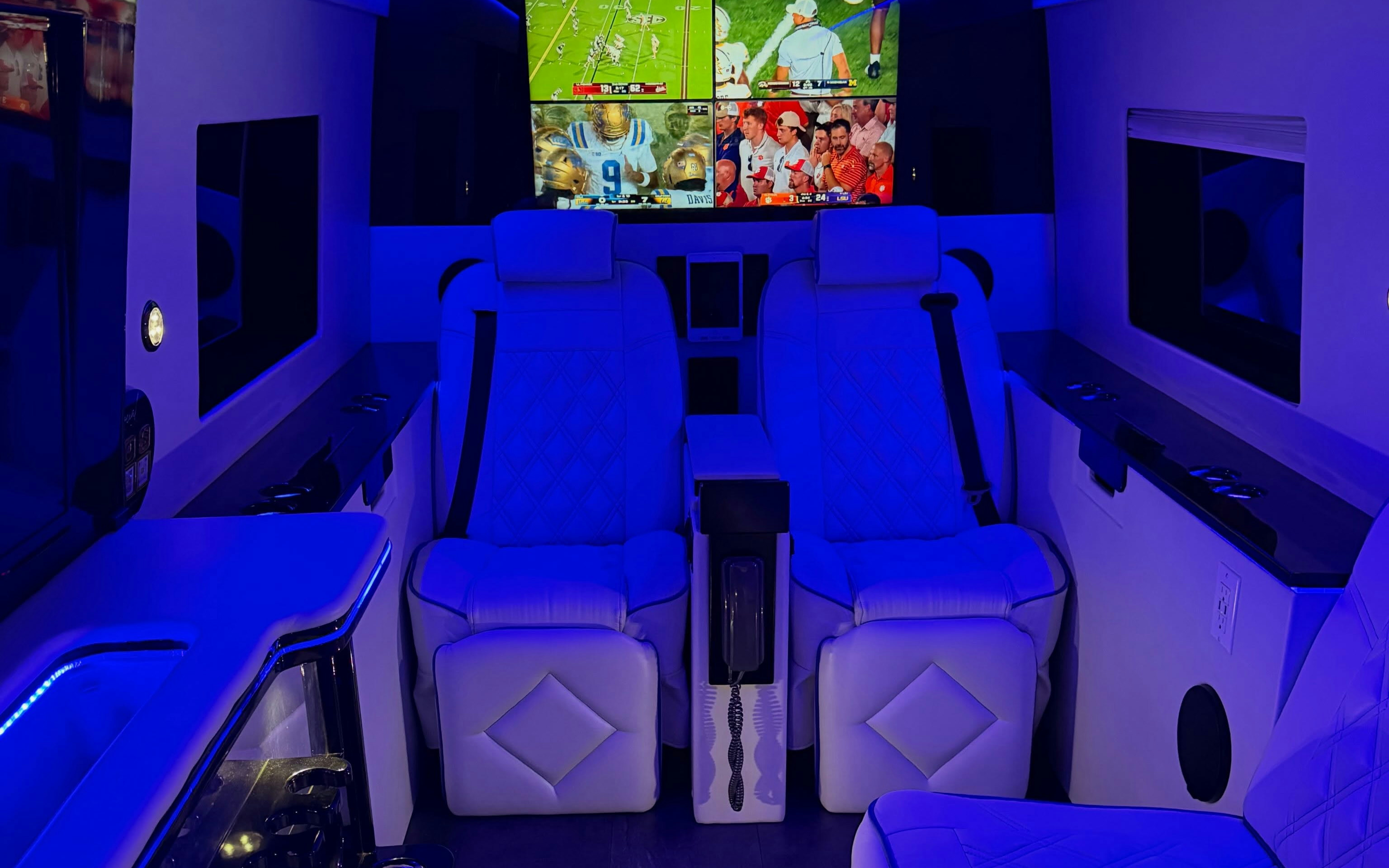 Luxury Sprinter van interior with white quilted leather captain chairs, blue LED lighting, and multiple TVs showing college football games