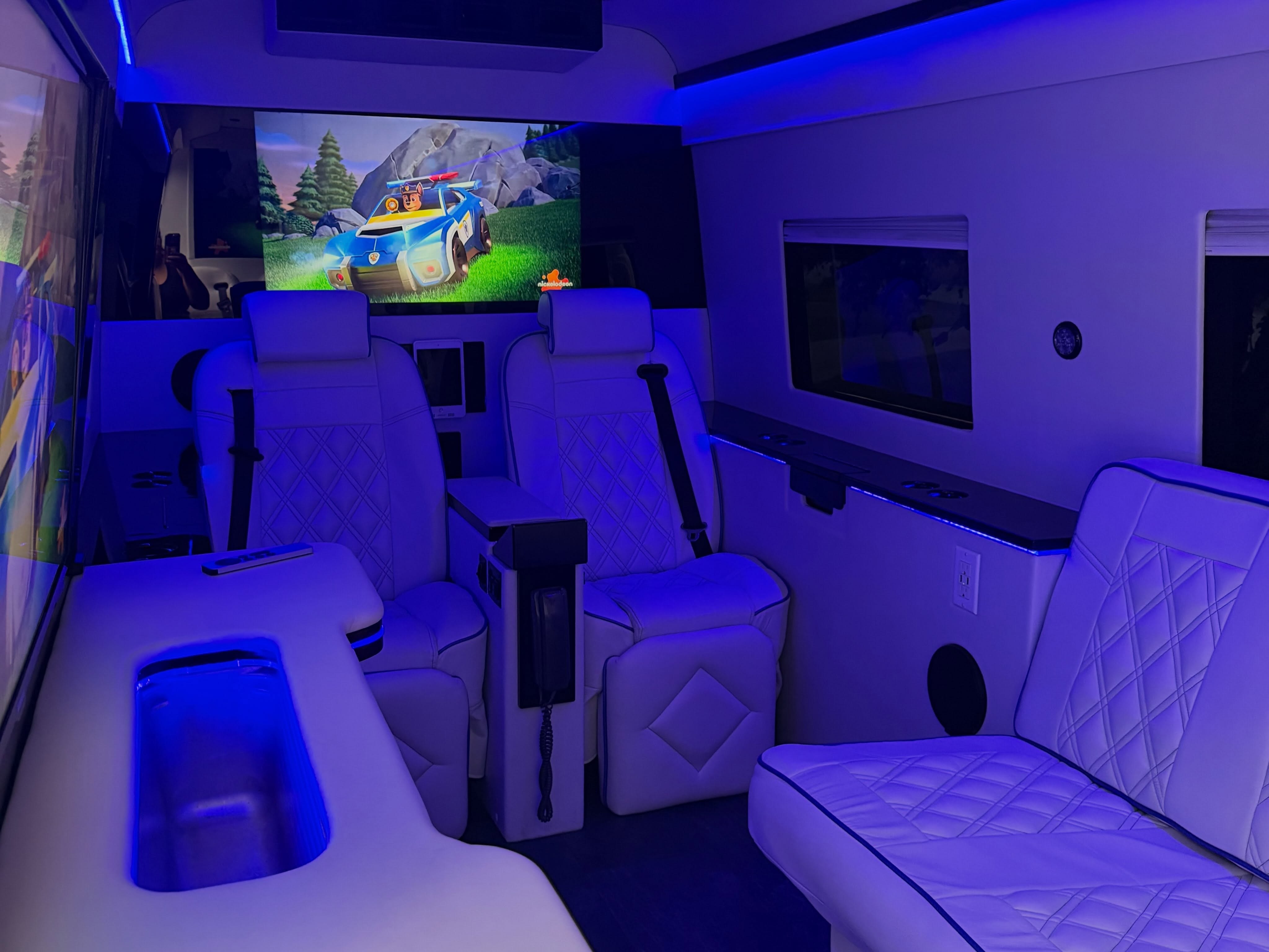 Luxury van interior with white quilted leather seats, blue LED lighting, and a TV screen showing a cartoon