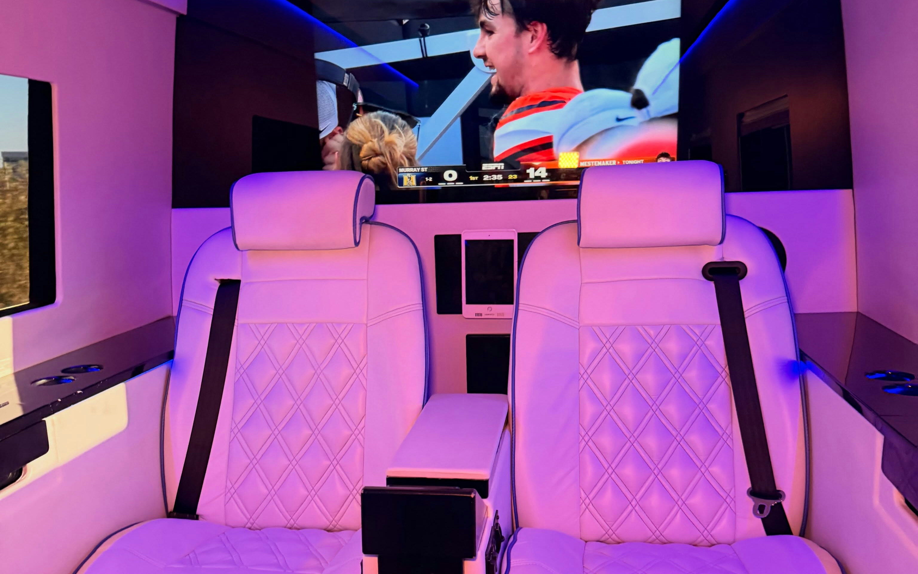 Luxury van interior with white quilted leather seats, purple ambient lighting, and a large TV screen showing an ESPN broadcast